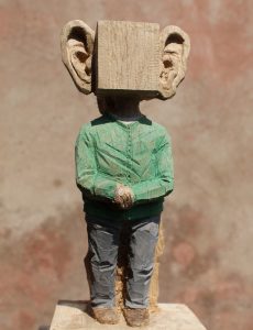 Ein stehender Mann auf Sockel vor ockerfarbenem Hintergrund. Er trägt ein grünes Hemd ind graue Hosen. Die Hände sind vor dem Buch gefaltet. Sein Kopf ist ein Würfel mit riesigen Ohren.