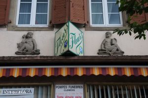 Zwei Sandsteinfiguren, dazwischen ein Schild mit der Aufschrift 'Fleurs'.