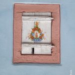 Ein geschlossener, weißer Fensterladen mit rosa bemaltem Sandsteingewänd. Ehemals ein Viehstall womöglich. Der Laden ist mit Blümchen bemalt.