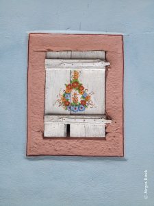 Ein geschlossener, weißer Fensterladen mit rosa bemaltem Sandsteingewänd. Ehemals ein Viehstall womöglich. Der Laden ist mit Blümchen bemalt.