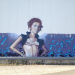 Ein Mädchen als kunstvolles Graffiti stützt die Arme in die Hüfte. Hintergrund türkis-graublaue Verzierung an kahler Betonwand