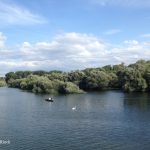 Graublauer Fluss in der unteren Bildhälfte, oben blauer Himmel und leichte Wolken, dazwischen viel Grün.