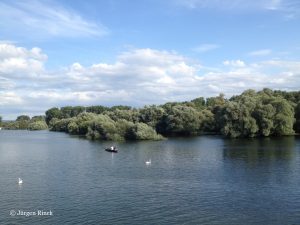 Graublauer Fluss in der unteren Bildhälfte, oben blauer Himmel und leichte Wolken, dazwischen viel Grün.
