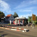 Über die Straße und eine Verkehrsinsel geblickt sieht man ein ovales Gebäude, Bäume und blauen Himmel