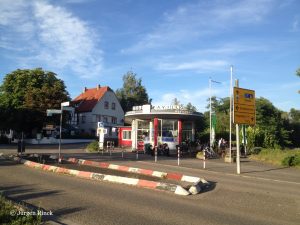 Über die Straße und eine Verkehrsinsel geblickt sieht man ein ovales Gebäude, Bäume und blauen Himmel