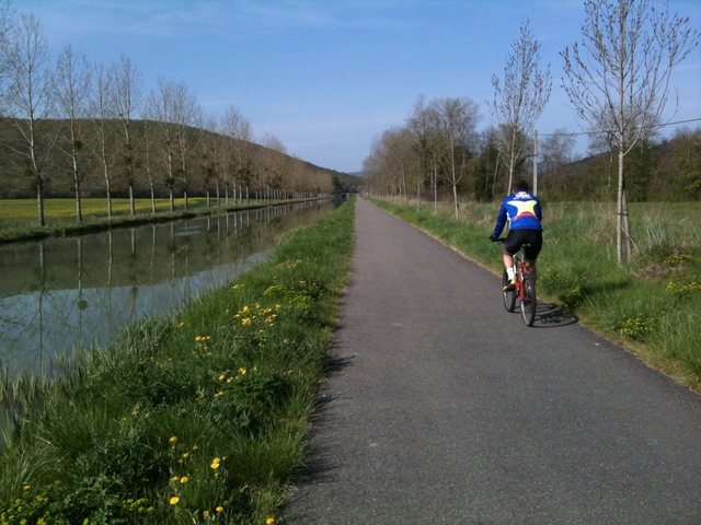 Radweg entlang eines Kanals. Frühling, noch jungknospende Pflanzen. Ein Rennradler auf dem schmalen geteerten Weg, der rechts vom Kanal verläuft.