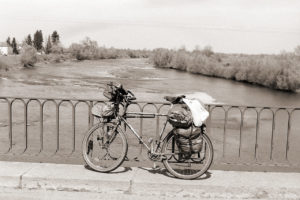 Schwarzweißbild eines Reiserads, das am Geländer einer Brücke über einen großen Fluss mit Stromschnellen lehnt