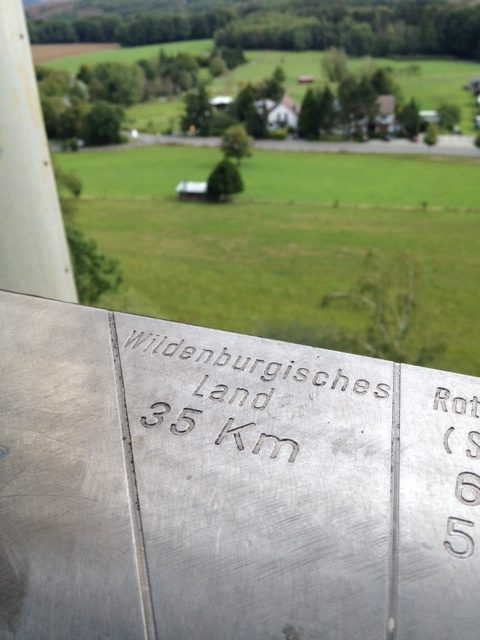 Aussicht vom Turm aus in das Wildenburgische Land, das laut der Anzeige auf dem Panoramazeiger im Vordergrund 35km entfernt ist. Der metallene Panoramazeiger ist im Vordergrund scharf gestellt, der Hintergrund, bestehend aus Wiesen, Streuobstbäume, eine Siedlung und im Hintergrund Wald, ist unscharf.