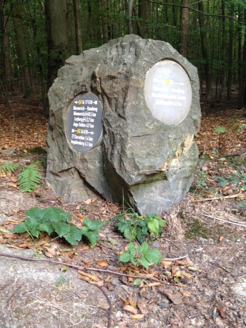 Kantiger Felsbrocken am Wegrand auf Waldboden, im Hintergrund Wald. Auf zwei Seiten des Steins zeigen Wegweiserschilder die Richtung an.
