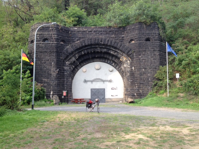 Der Tunneleingang zum Theater im Tunnel unter der Erpeler Ley ist ein hoher, aus dunklem Stein gebauter, symetrischer Bau. Beidseitig turmähnlich, in der Mitte ein Bogen, der Eingang selbst zugemauert, doch in der Mitte des Rundbogen ist eine graue Türöffnung zu sehen. in der Bildmitte, vor der grauen Tür das Reiserad. Rechts eine Europafahne auf Fahnenstange, links die Deutschlandfahne. Alles umgeben von Bäumen, im Vordergrund der Radweg.