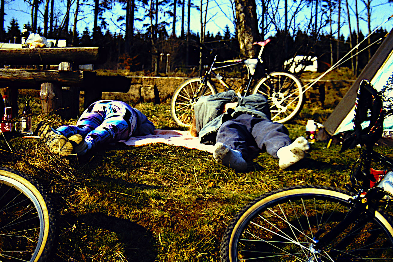 Eine Picknickwiese, auf der zwei Radler schlafen. Davor liegen Fahrräder auf dem Boden, im Hintergrund lehnt ein Fahrrad an einem Baum. Die Socken eines der Radler sind nicht sehr sauber.