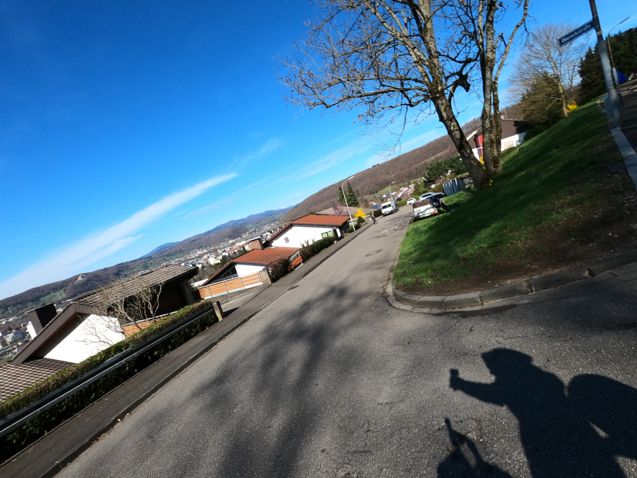 Schräg aufgenommenes Foto, Horizont oben rechts nach unten links. Eine Frühlingsbild mit grün und blauem Himmel, keimenden Bäumen. Auf dem Teer der Straße der Schatten eines fotografierenden Reiseradlers.