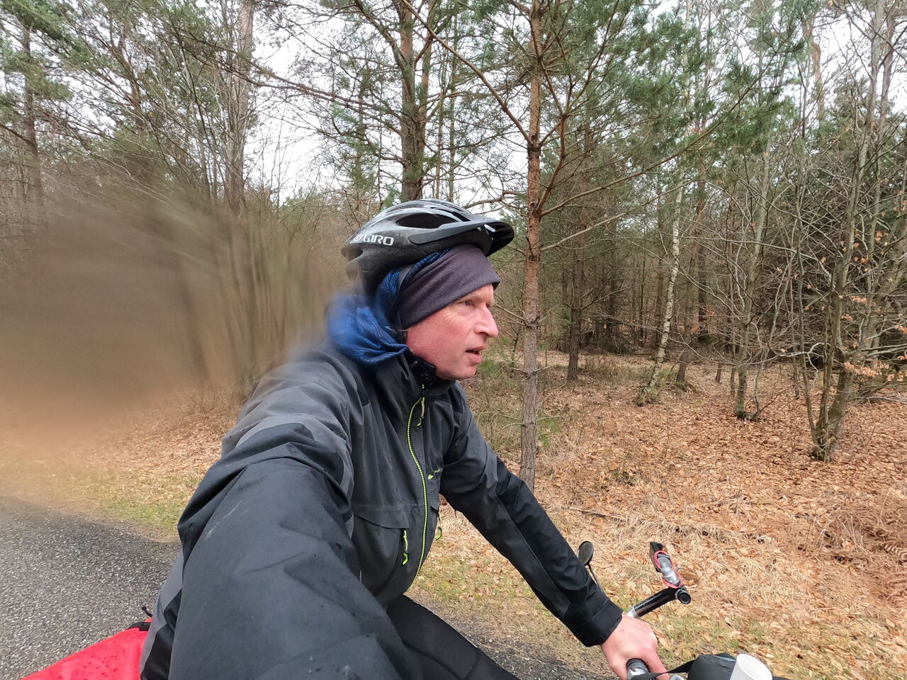Mensch mit Regenkleidung und Fahrradhelm auf dem Fahrrad von der Seite sich selbst fotografierend. Im Hintergrund ein lichtes Wäldchen, das noch kaum Laub trägt.
