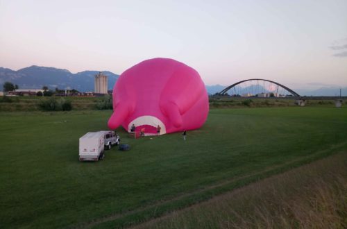 Pink farbener Heißluftballon vor einer Brücke. Der Ballon liegt noch und winzig steht daneben ein Auto mit Anhänger