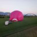 Pink farbener Heißluftballon vor einer Brücke. Der Ballon liegt noch und winzig steht daneben ein Auto mit Anhänger