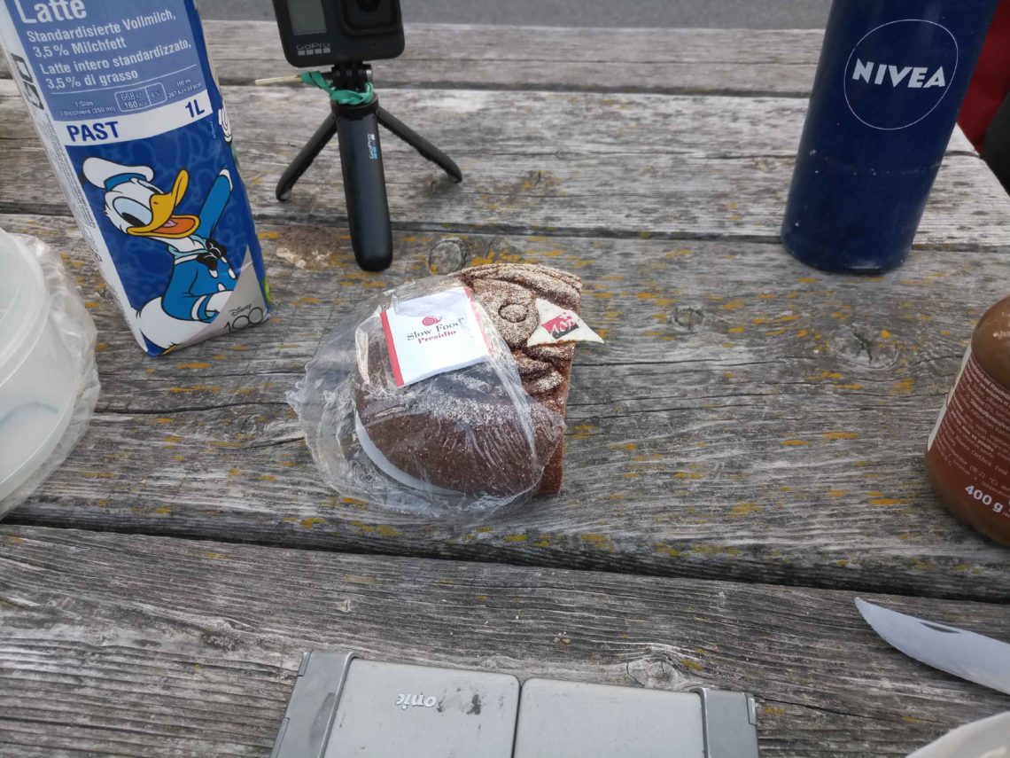 Picknicktisch mit einer Flasche Milch, einem runden, dunklen Brot in Plastikfolie, halb ausgepackt und dem Stativ einer Actionkamera