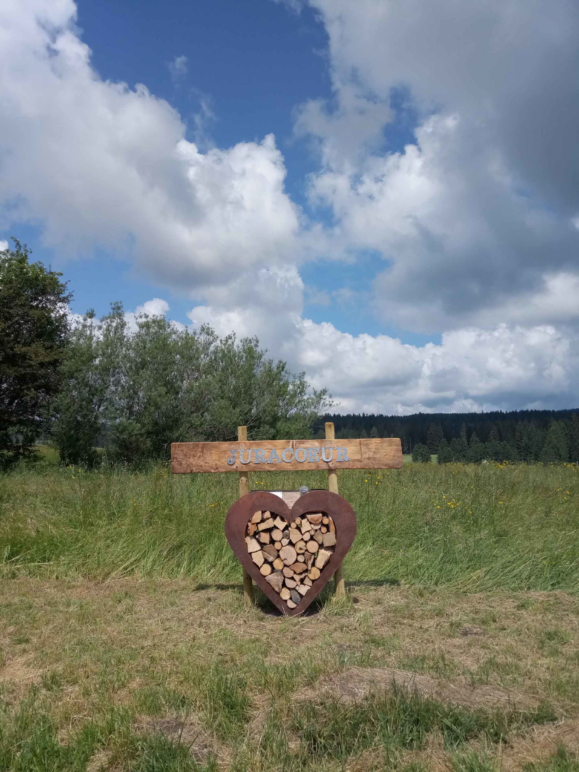 Ein Herz aus Holzscheiten, gefasst in rostigem Eisen weist auf eine Pension hin. Quellwolken stehen am Himmel über der Bergszenerie.