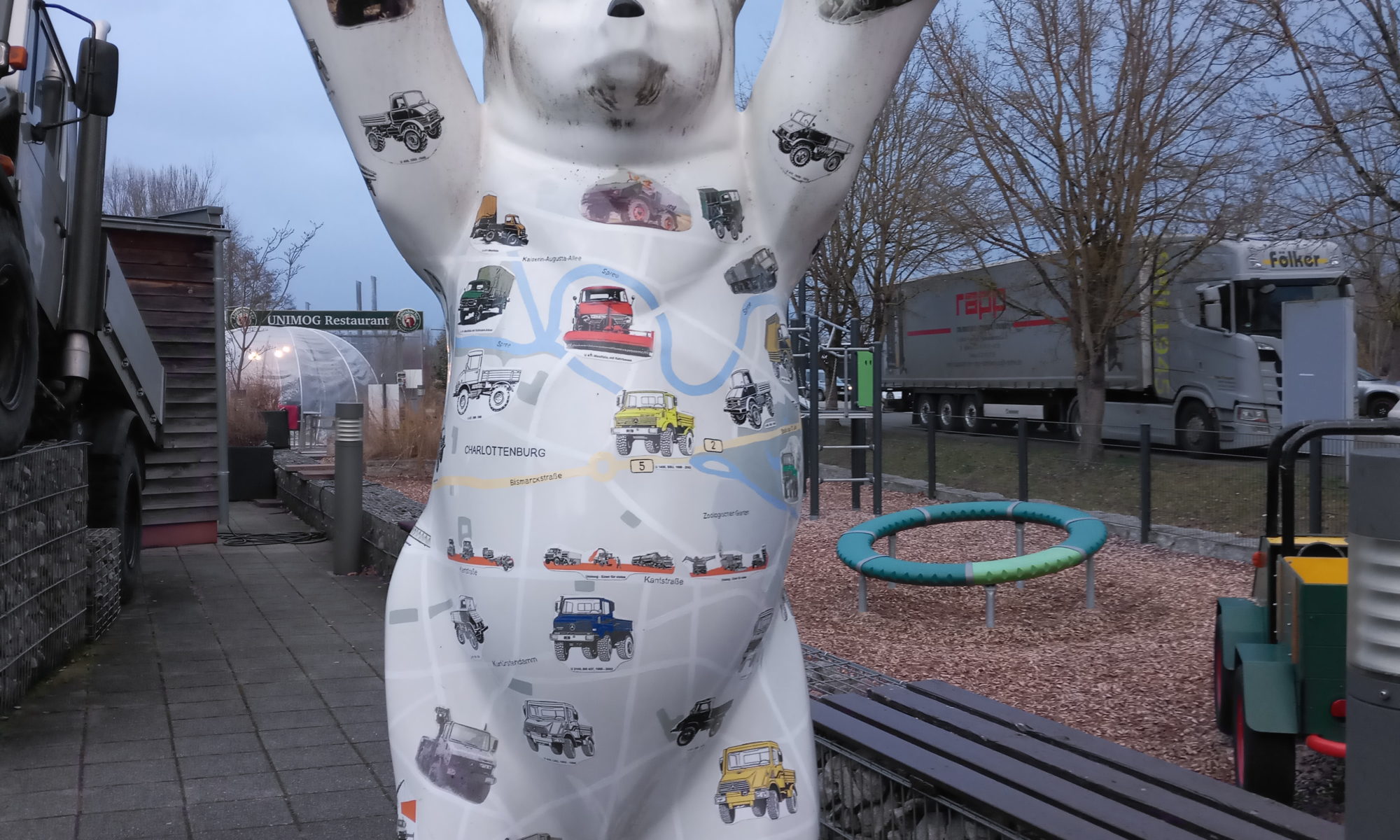 Hochkantbild einer Bärenskulptur aus Polyester. Der weiße Bär steht auf zwei Beinen und Hält die Ortsschilder von Berlin und das Ortsausgangsschild von Berlin, durchgestrichen mit Hinweis auf Gaggenau in 700 km nach oben. Er ist beklebt mit zahlreichen bunten Stickern von Unimog-Fahrzeugen. Der Hintergrund aus Pflastersteinen, einer Sitzbank und einem Spielplatz und Gemäuer lässt eine innerstädtische Position vermuten vorm Protal des Unimogmuseums ind Gaggenau.