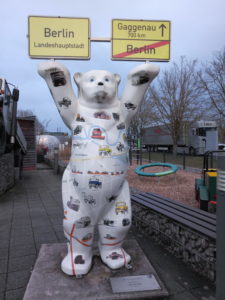 Hochkanbild einer Bärenskulptur aus Polyester. Der weiße Bär steht auf zwei Beinen und Hält die Ortsschilder von Berlin und das Ortsausgangsschild von Gaggenau, durchgestrichen mit Hinweis auf Berlin nach oben. Er ist beklebt mit zahlreichen bunten Stickern von Unimog-Fahrzeugen. Der Hintergrund aus Pflastersteinen, einer Sitzbank und einem Spielplatz und Gemäuer lässt eine innerstädtische Position vermuten vorm Protal des Unimogmuseums ind Gaggenau.