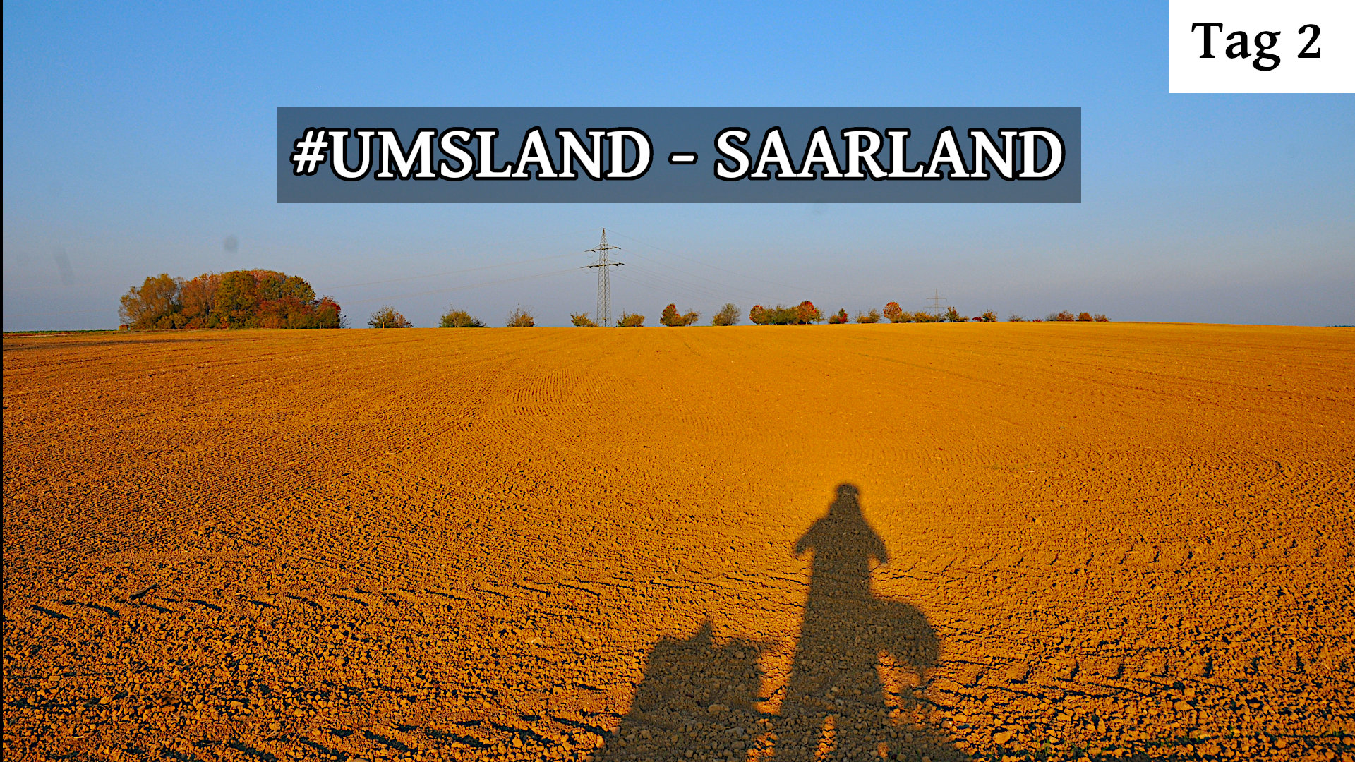 Blick auf ein weites, gelbliches, abgeerntetes Feld mit Baumreihe im Hintergrund. Ein langer Schatten eines Radlers mit viel Gepäck am Rad fällt auf das Feld. Der Schriftzug UmsLand Saarland prangt im Blau des Himmels. Oben rechts steht geschrieben Tag 2.