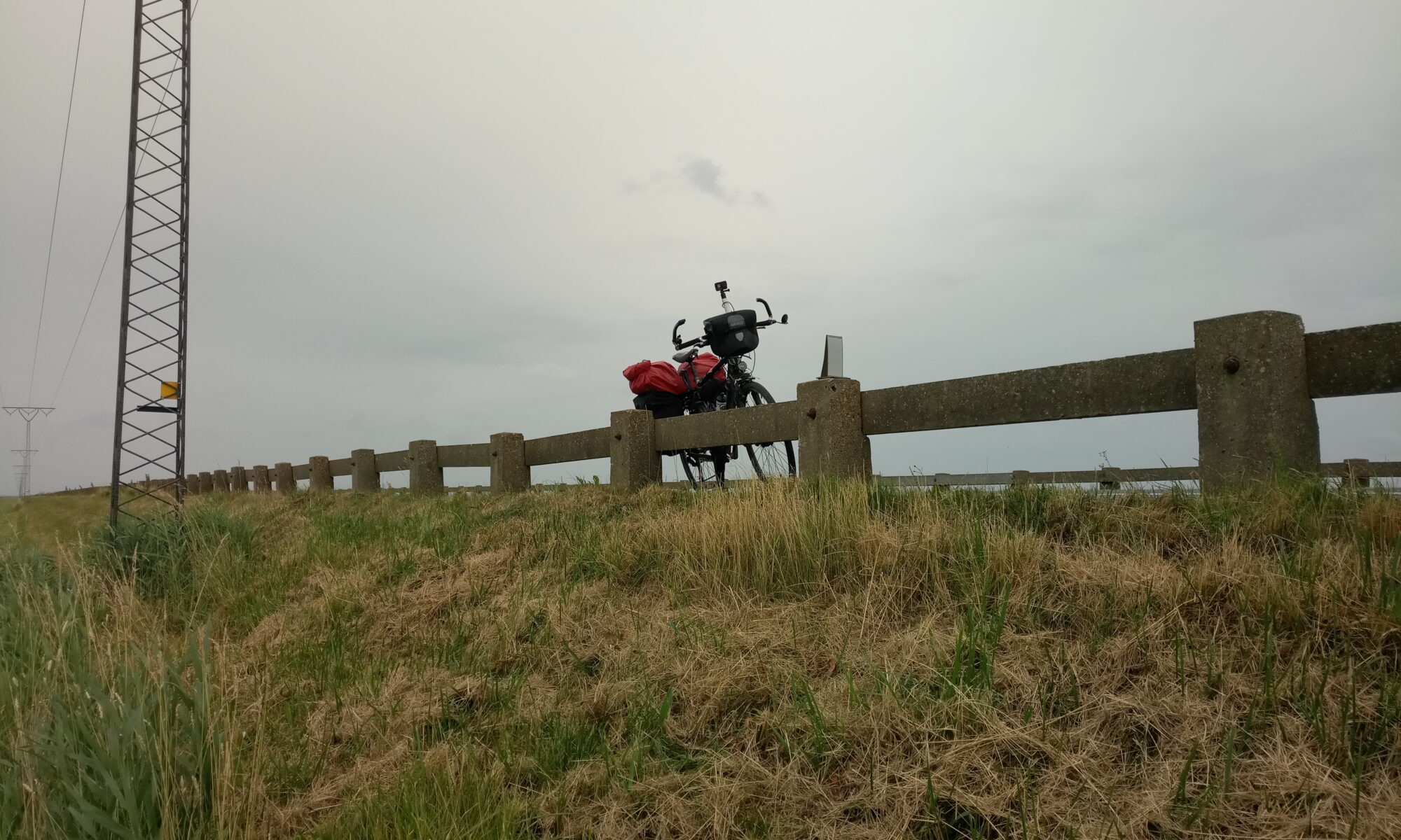 Am Fuß eines Deiches etwas nach oben gerichtet folgt der Blick einer Leitplanke, an der ein Fahrrad angelehnt ist. Regnerisch wirkt der Himmel.