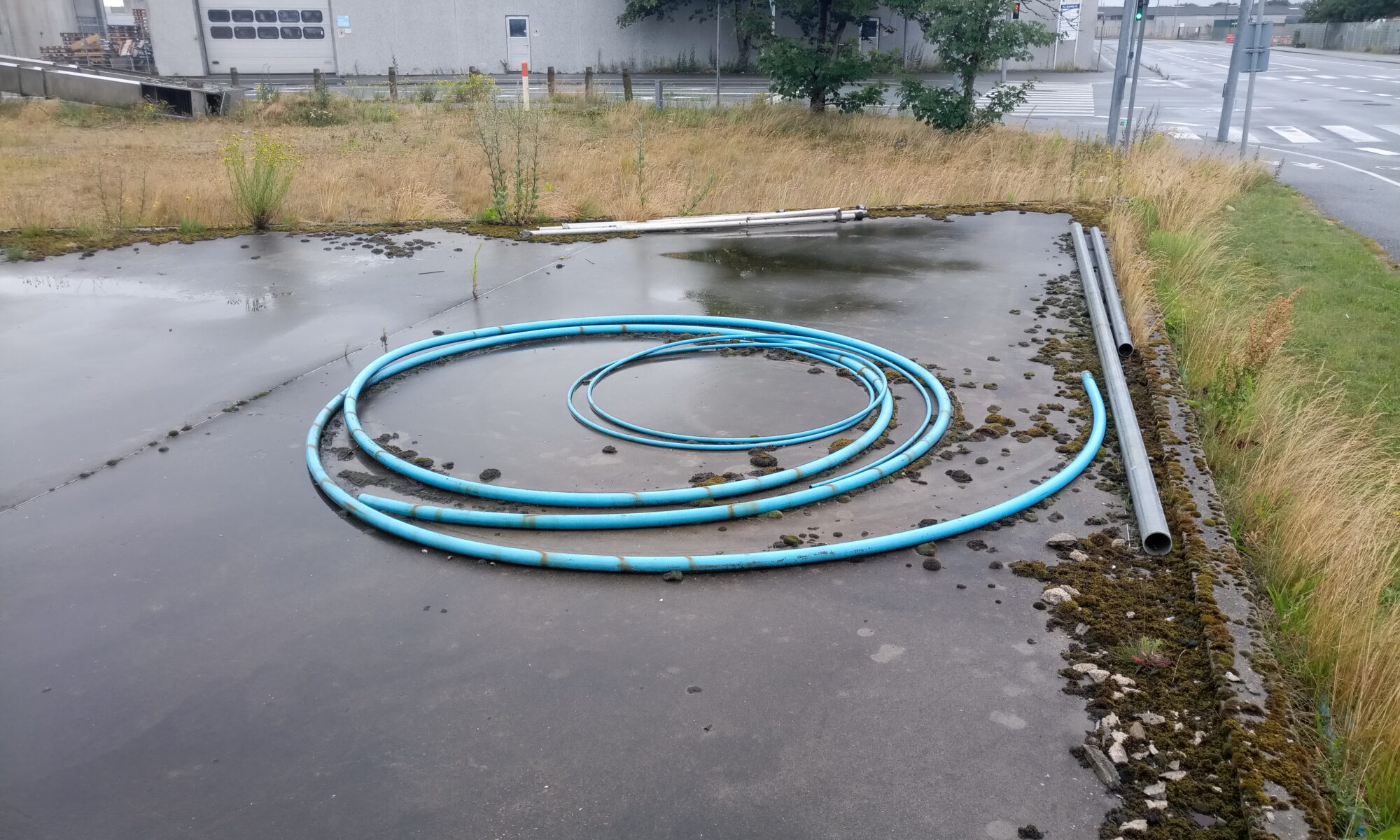 Auf einem verregneten Industriegelände liegen türkisfarbene Schläuche. Im Hintergrund ein Industriebauwerk und kleine Sträucher.