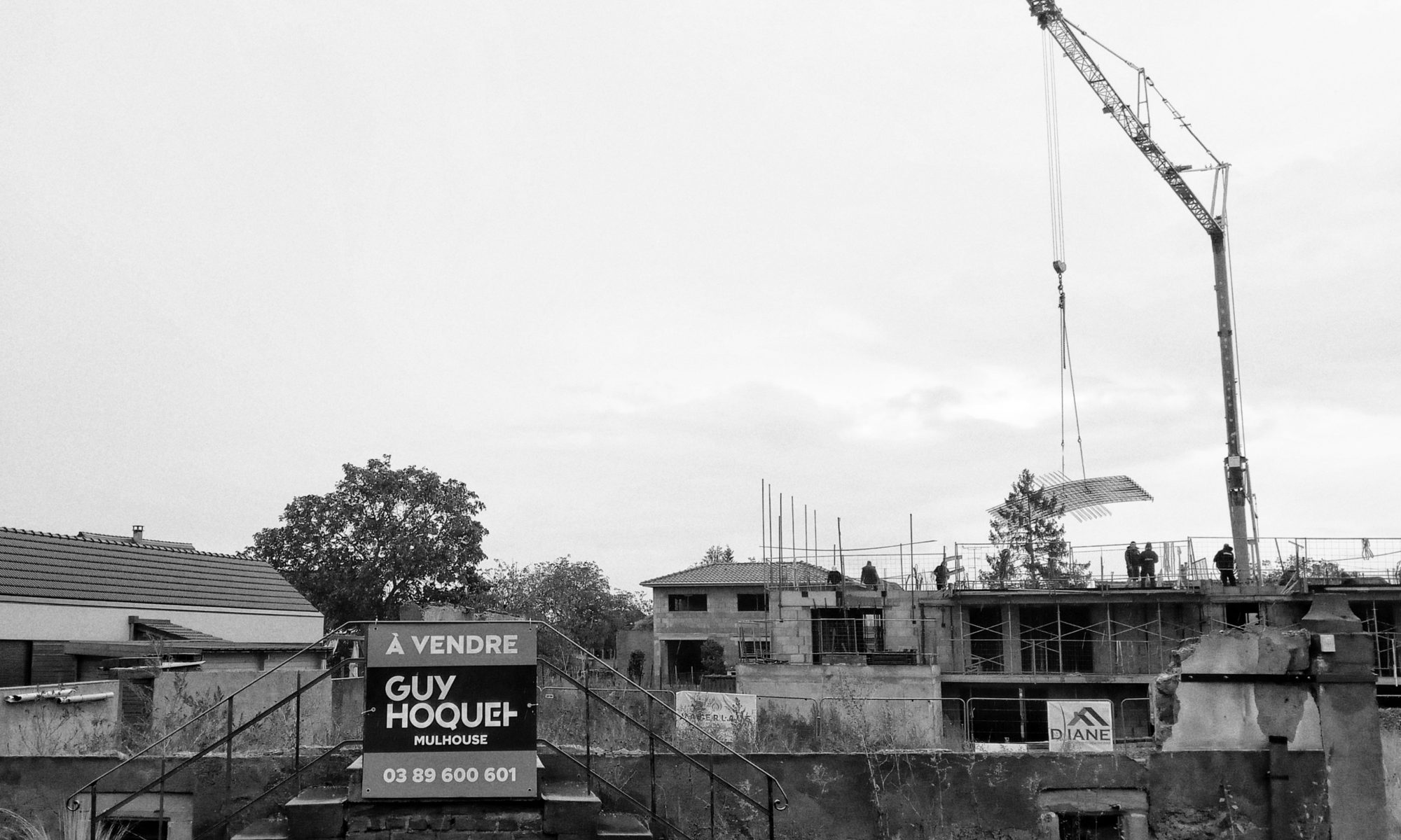 Schwarzweißbild einer Abrissbaustelle am dörflichen Straßenrand. Dominant steht im Vordergrund links der Bildmitte noch eine zweiläufige Hauseingangstreppe, während dahinter schon neue Fundamente gelegt werden. Ein Baukran ist rechts im düsteren Bild zu sehen.