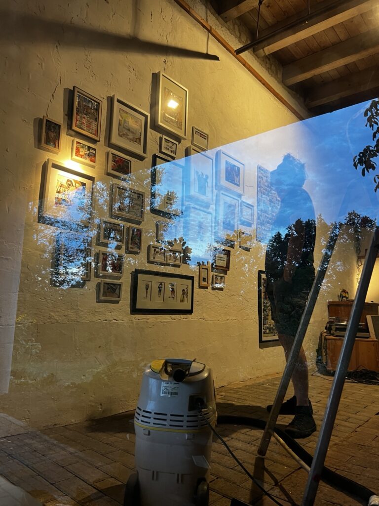 Blick durch die spiegelnde Scheibe auf ein Schattenreich iluminiertes Atelier in warmem Farbton. Ein Mann steht neben einer Leiter und einem Staubsauger und bestaunt eine Wand mit etwa dreißig unterschiedlich großen Bildern in Petersburger Hängung.